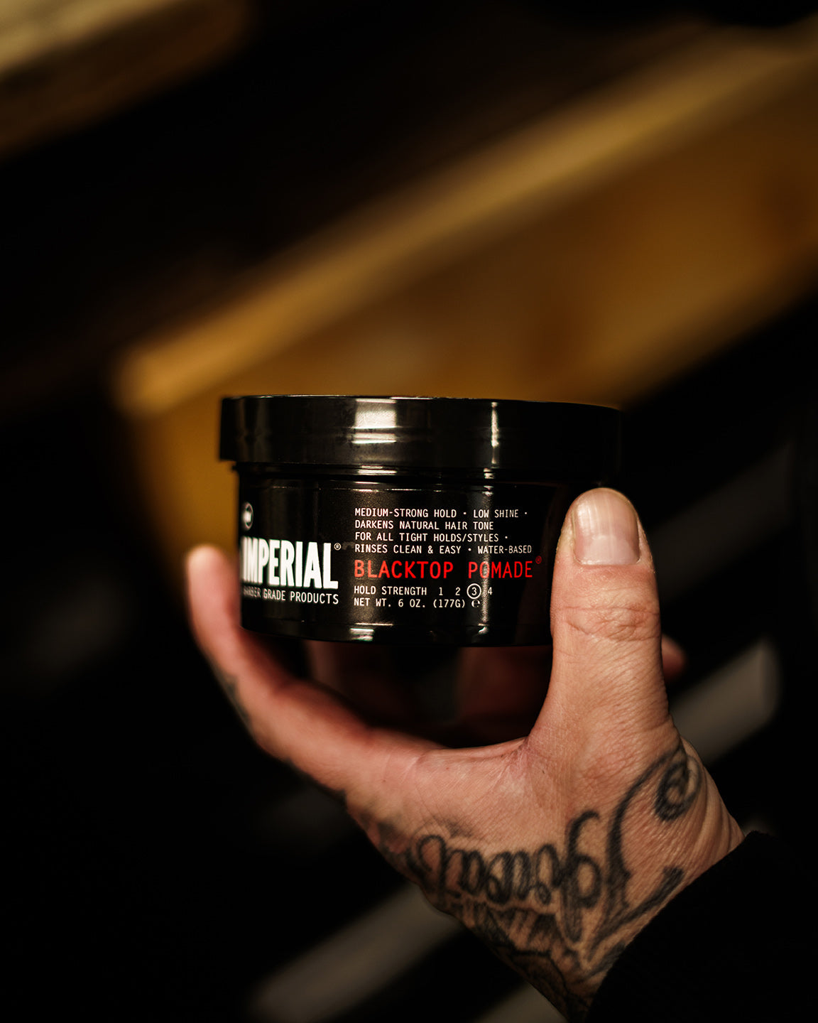 A barber's tattooed hand holding Blacktop Pomade